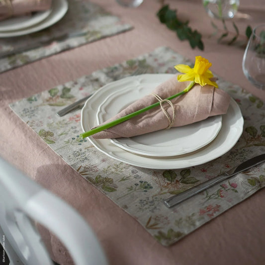 En dukning med brunrosa duk, blommiga bordstabletter och matchande tygservetter, där en påsklilja finns inlindad i ett natursnöre runt servetten.