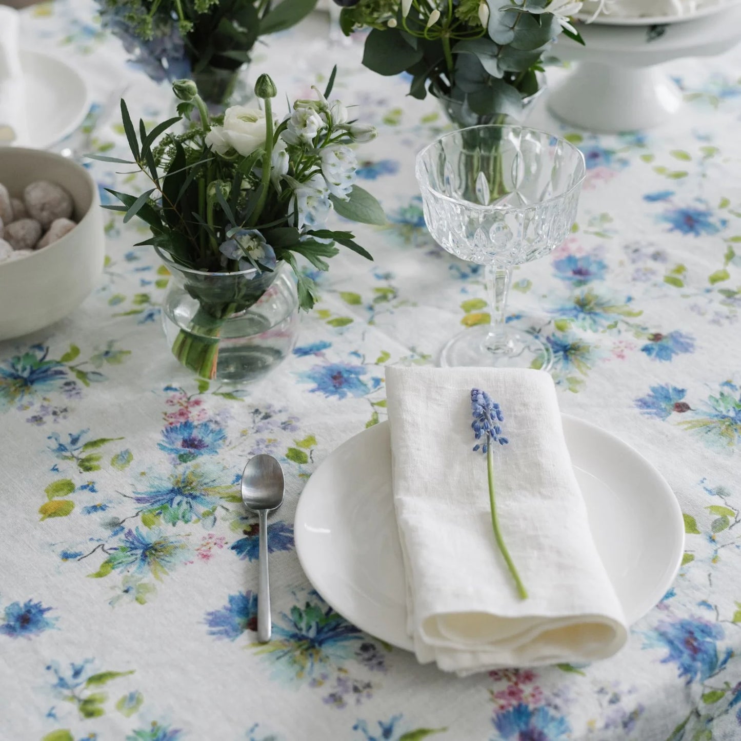 En vårig och romantisk dukning med en blåvit blommig duk och vit linneservett med en blå blomma på den vikta servetten. 