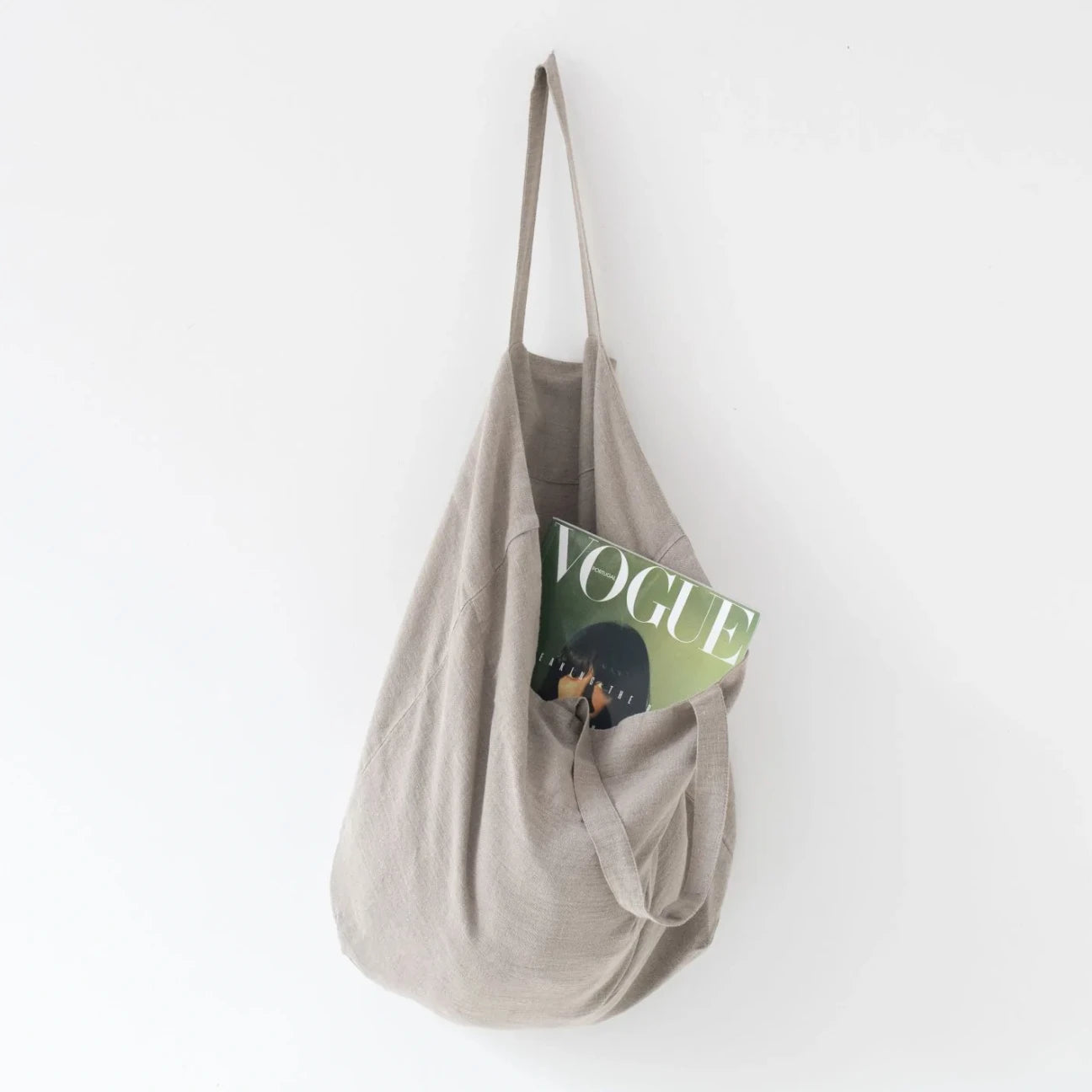 Medelstor tygväska i naturell linne som hänger på en krok med sitt ena handtag. Väskan har formen av en rektangel med medellånga handtag. Från väskan skymtar en Vouge tidning. 