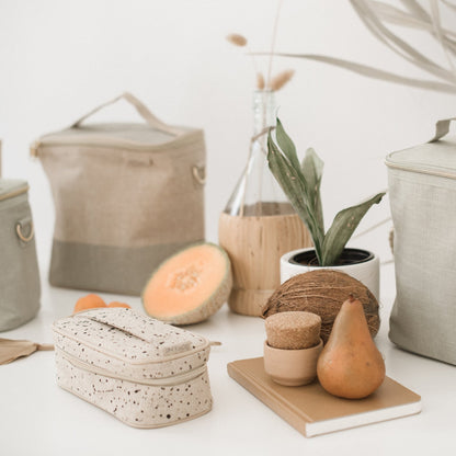 En liten rektangulär necessär och stora lunchväskor i bakgrunden i naturell linne med detaljer står på ett bord.