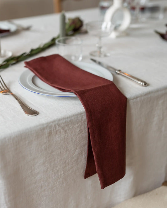En lång röd linneservett ligger på en vit tallrik på ett dukat bord. 
