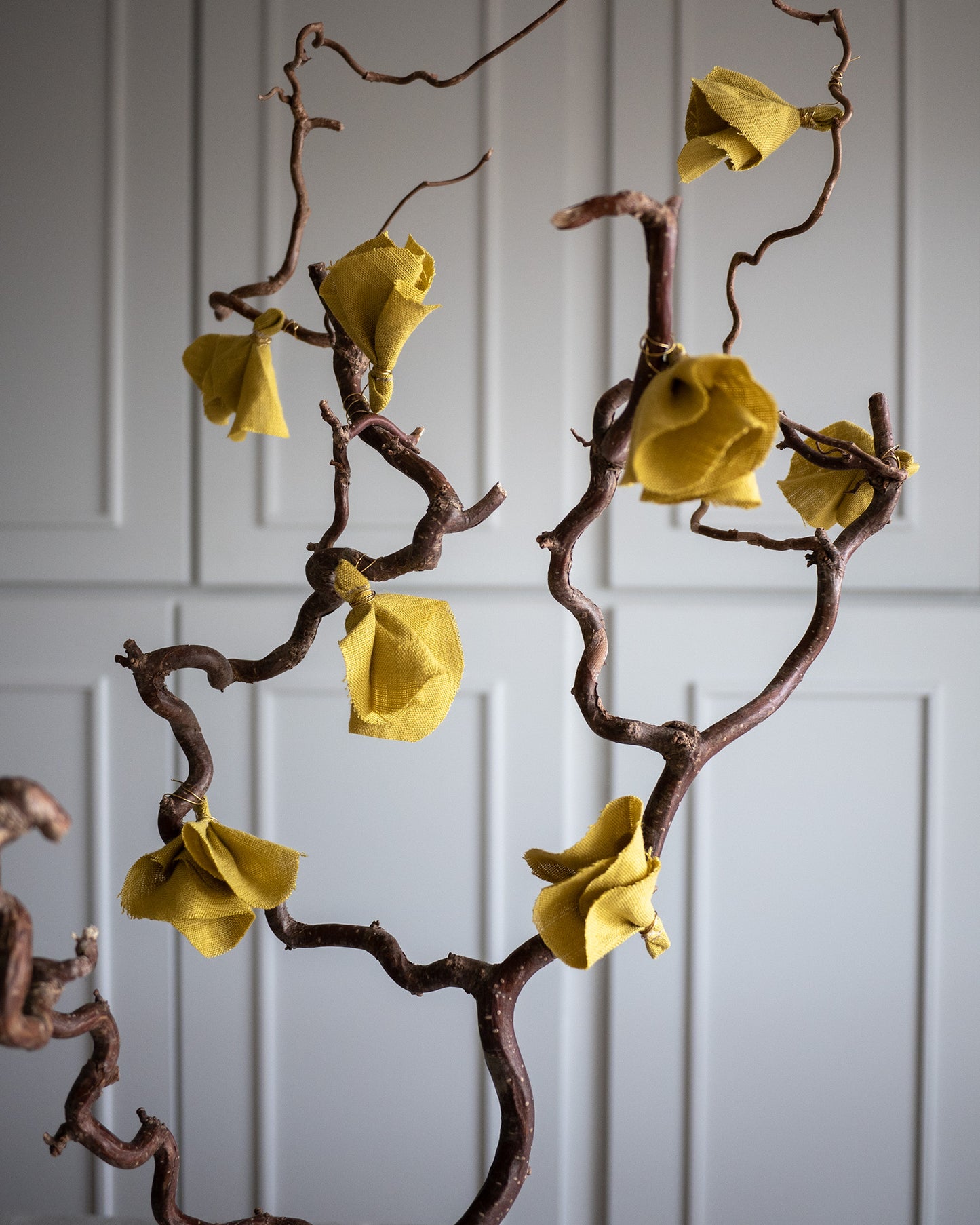 Åtta stycken små tygblommor i gult lin hänger på en trollhassel gren. 