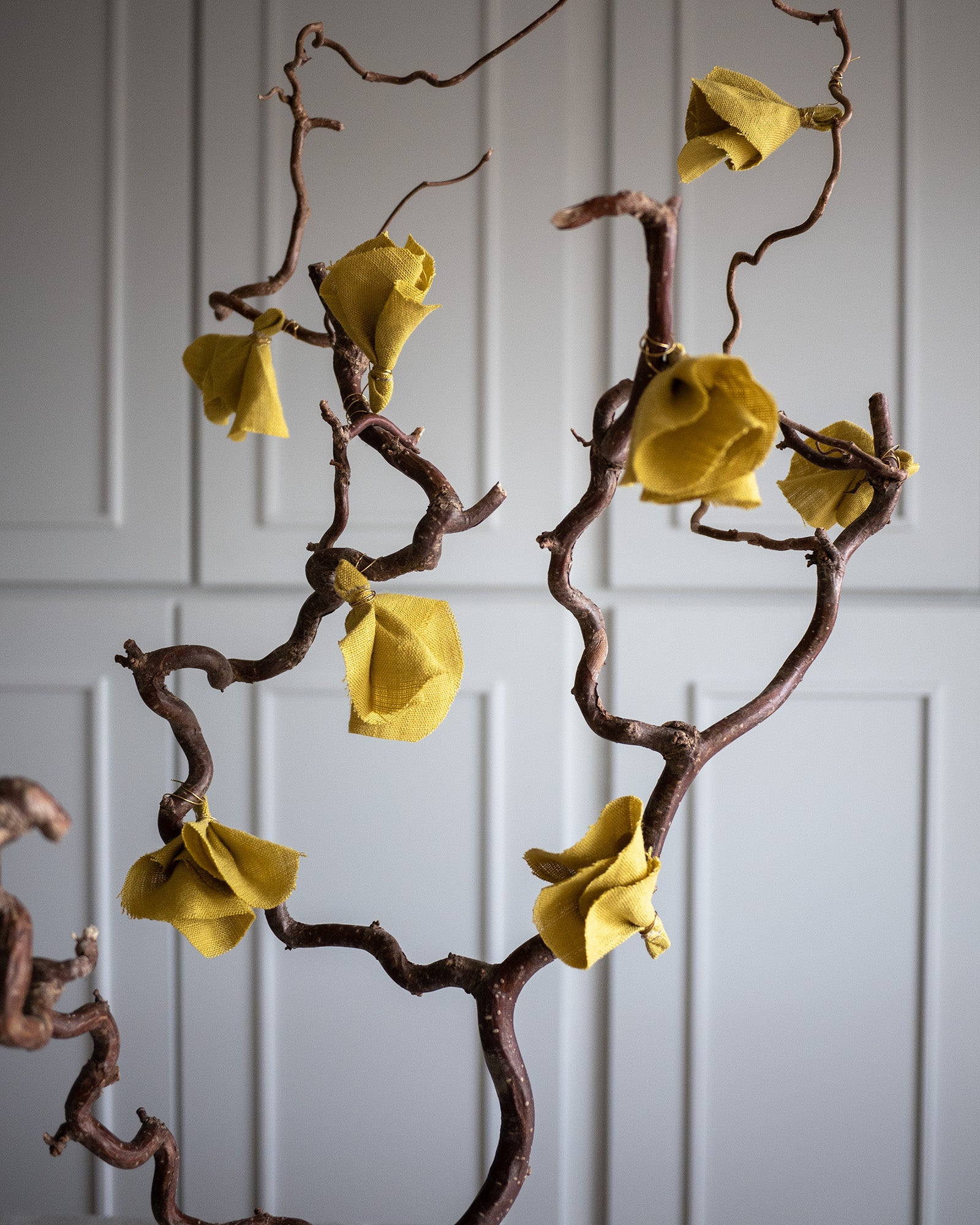 Åtta stycken små tygblommor i gult lin hänger på en trollhassel gren. 